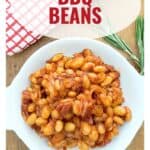 A bowl of BBQ beans on a wooden table with a red checkered towel and rosemary sprigs nearby.