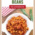 A white plate of BBQ beans on a wooden table with a red-checked towel and fresh rosemary nearby.