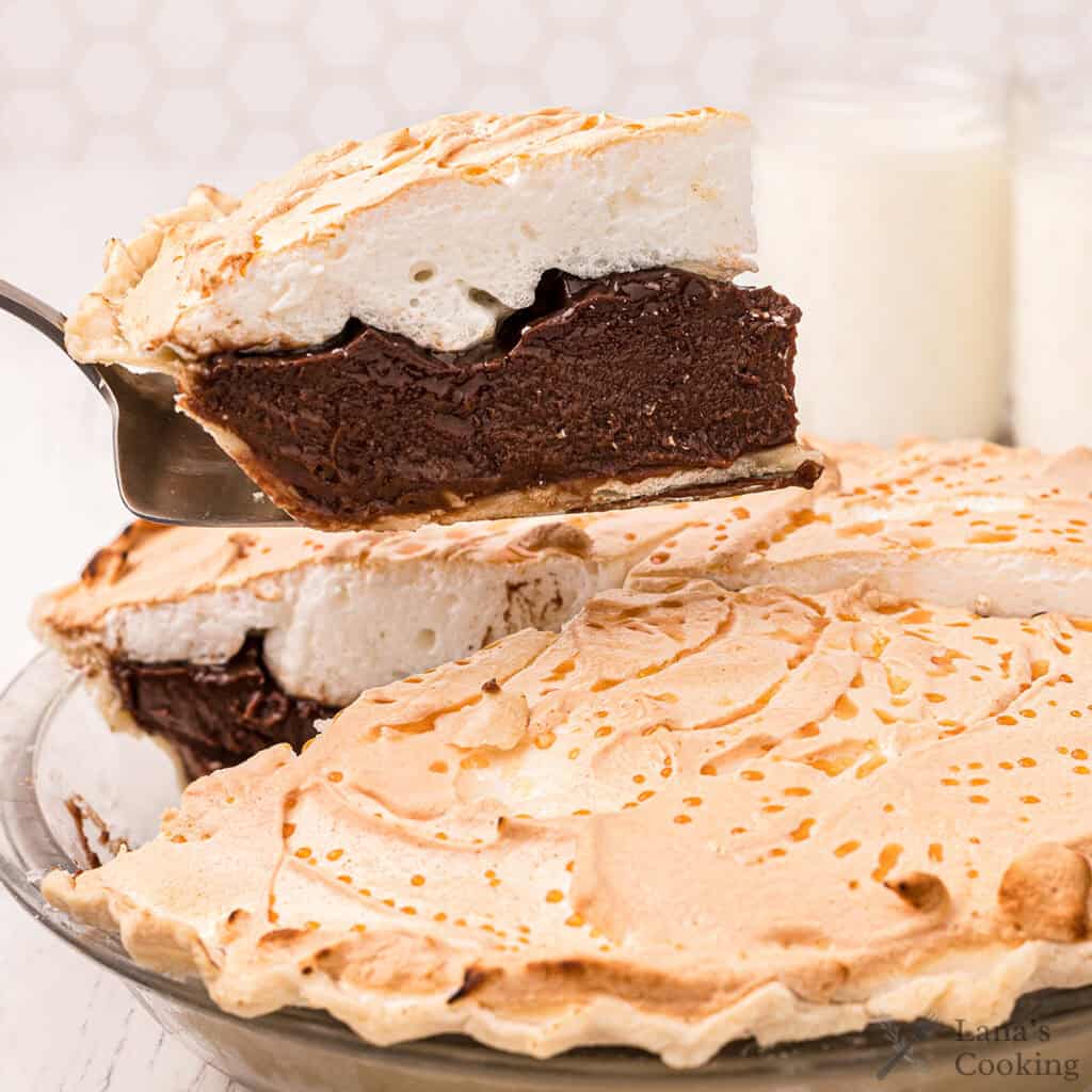 A slice of chocolate meringue pie is being lifted from a whole pie on a glass plate.