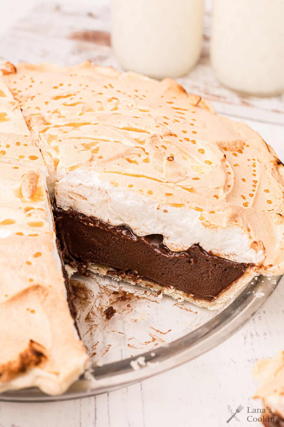 Chocolate pie with a layer of meringue topping, missing one slice, on a glass pie plate on a white table.