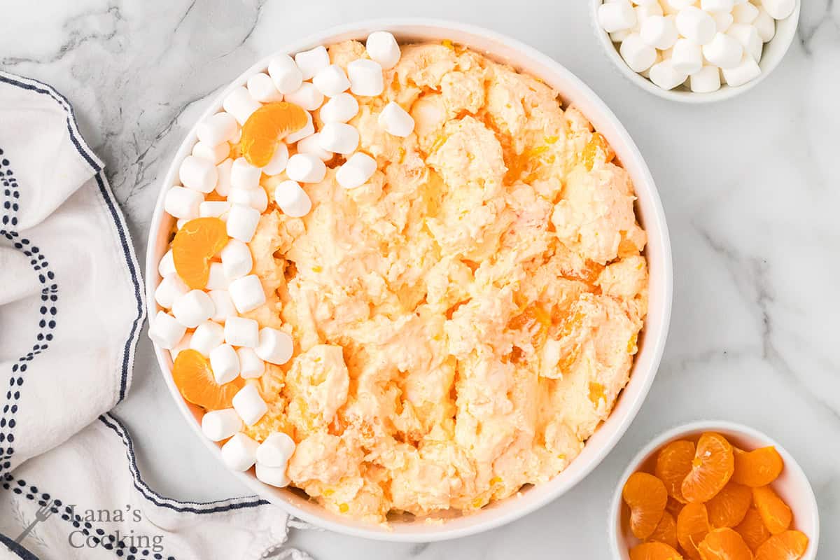 A bowl of orange fluff salad with mini marshmallows and mandarin orange slices on top, next to bowls of each.