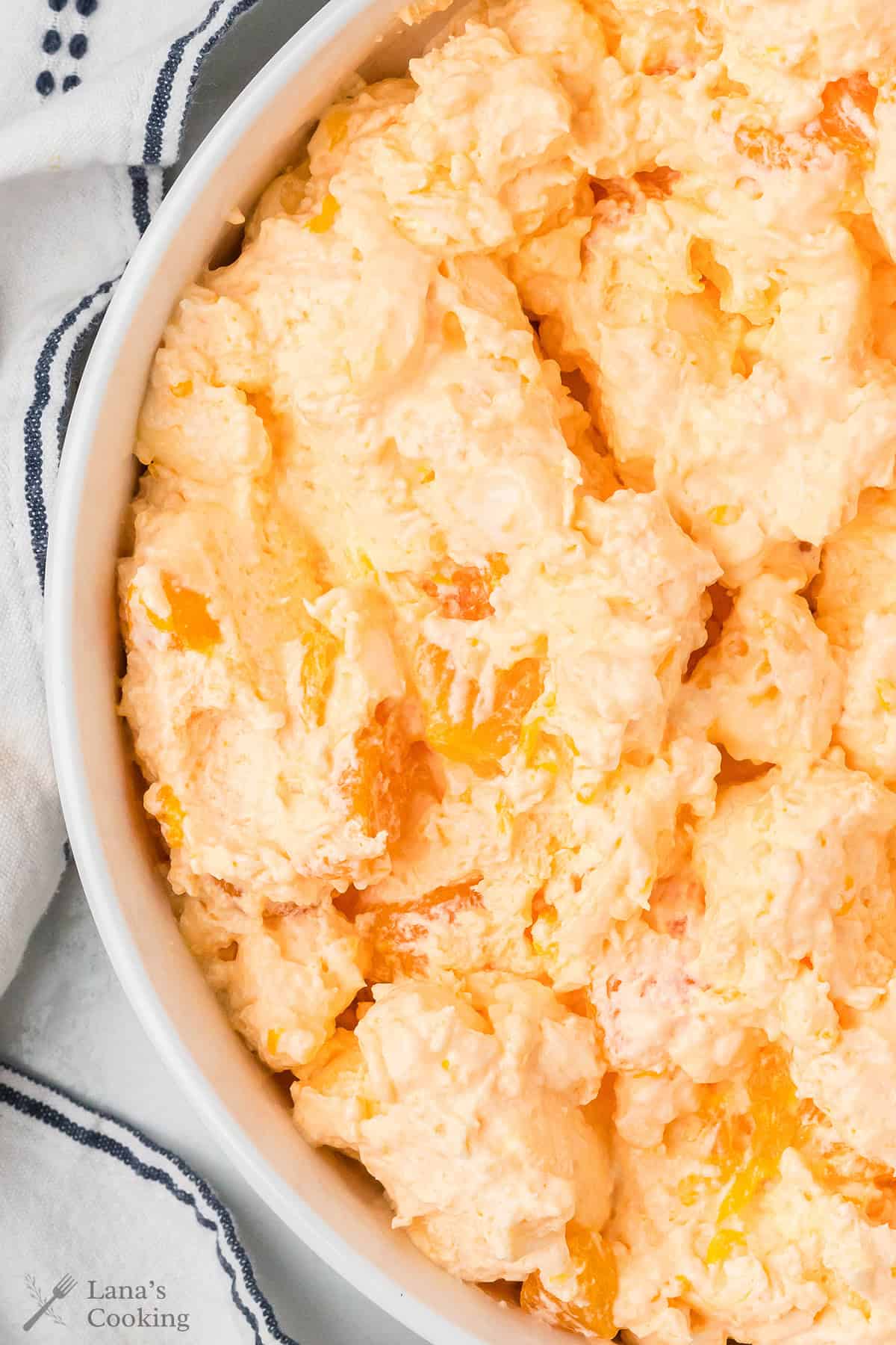 A close-up of a creamy orange fluff salad with mandarins in a white dish on a striped towel.