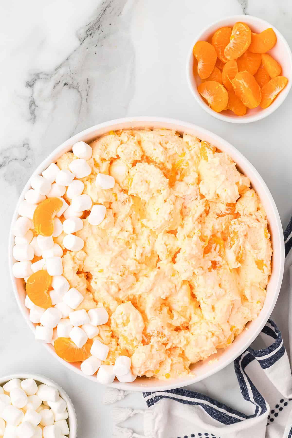 Large bowl of orange fluff salad with marshmallows and mandarin oranges, surrounded by extra toppings.