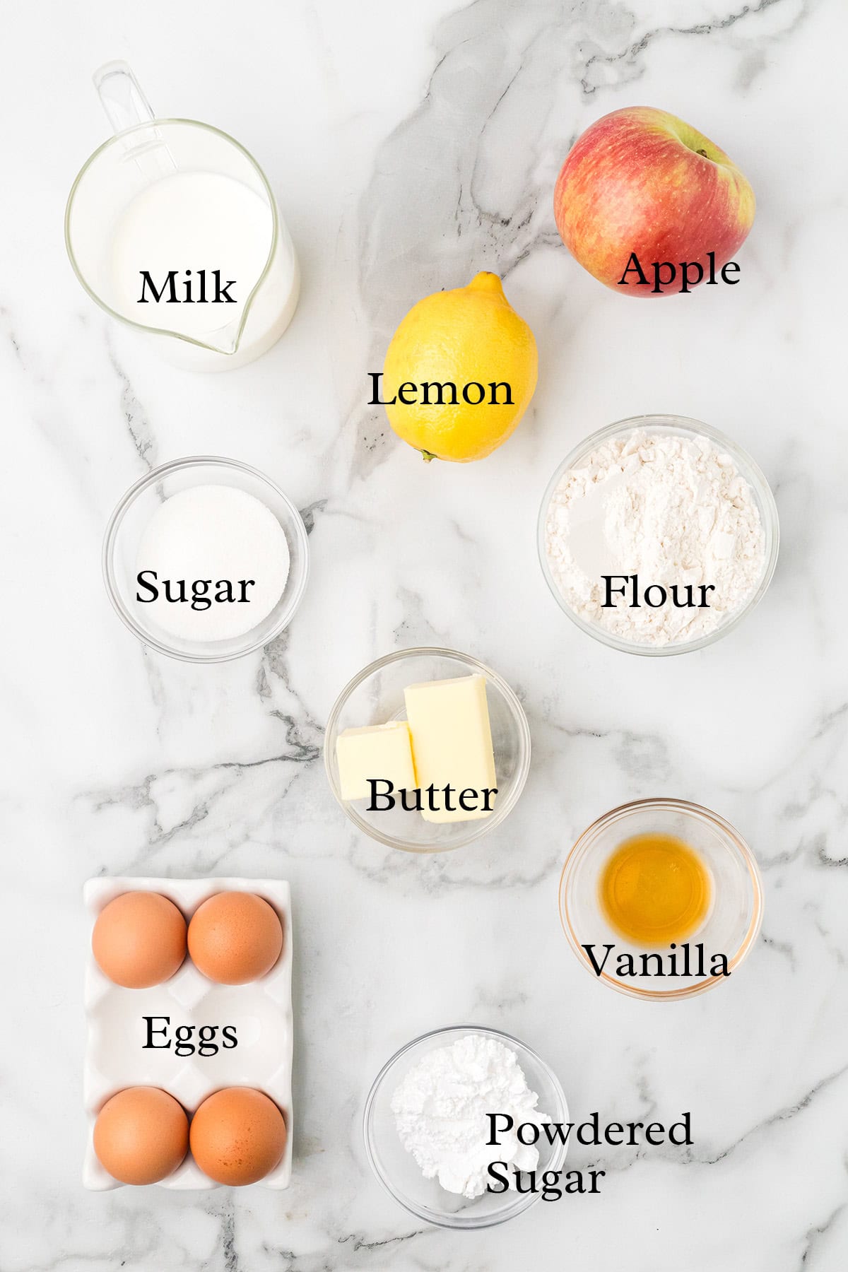 Top-down view of labeled baking ingredients: milk, apple, lemon, sugar, flour, butter, vanilla, eggs, powdered sugar.