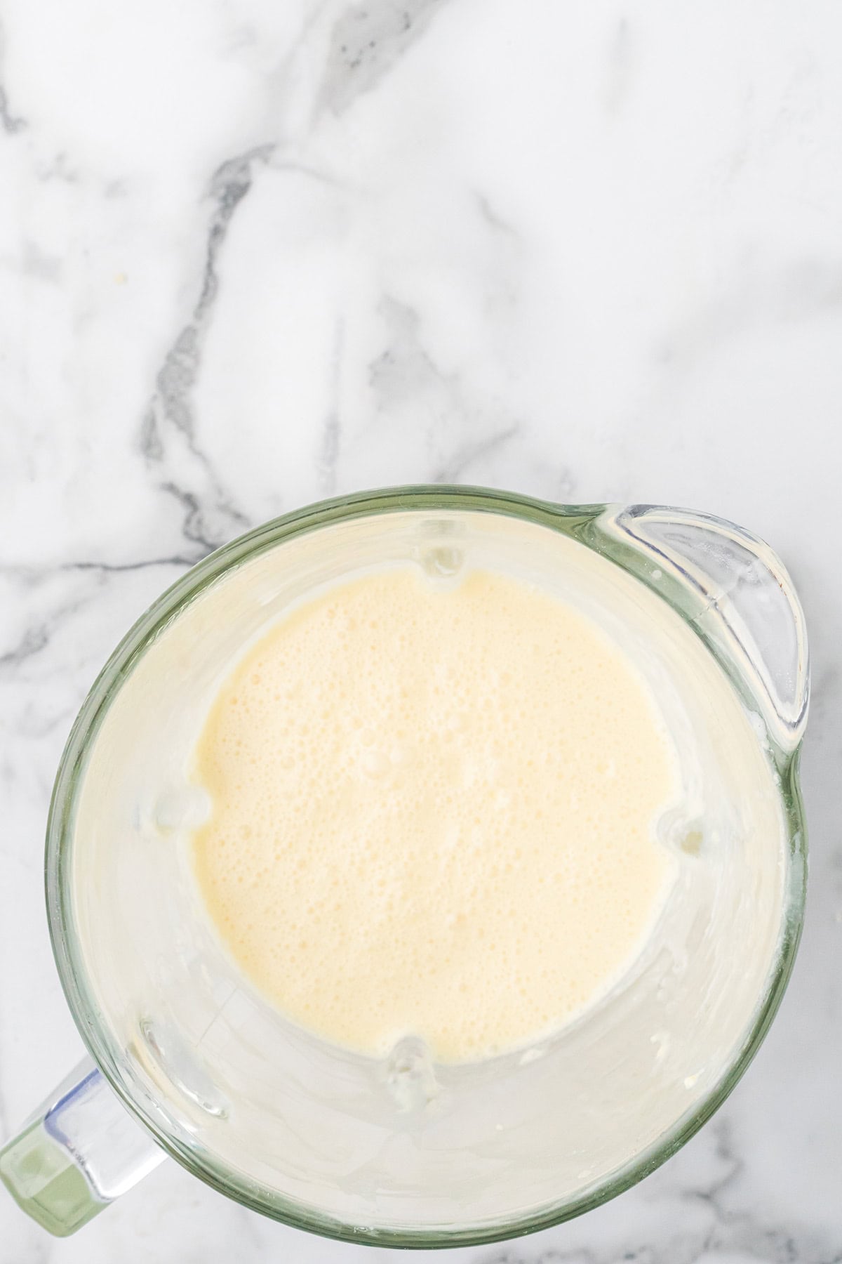 Top view of a glass blender filled with a creamy, pale yellow smoothie on a marble countertop.