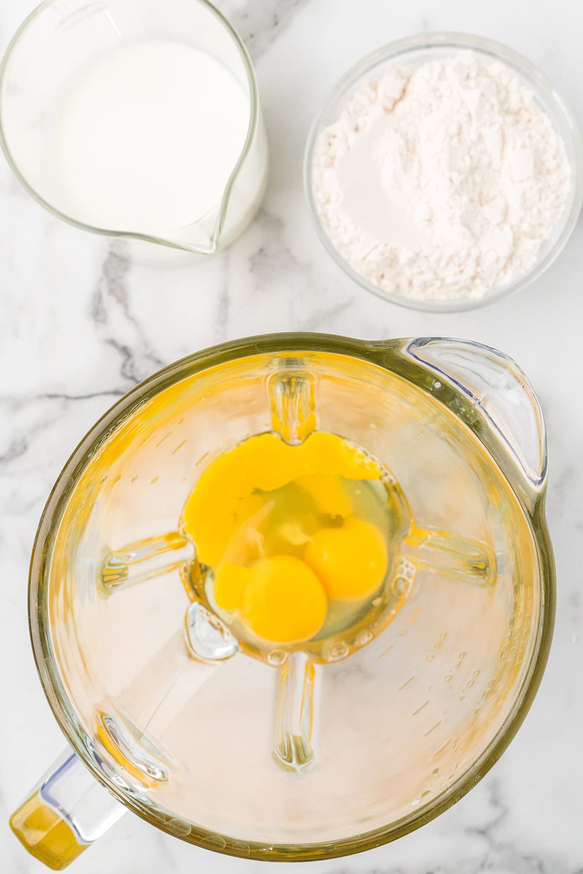 Top view of eggs in a blender, with a bowl of flour and a measuring cup of milk on a marble surface.