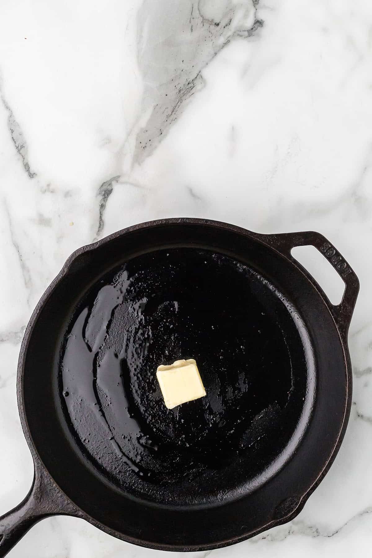 A pat of butter melting in a black cast iron skillet on a white marble countertop.