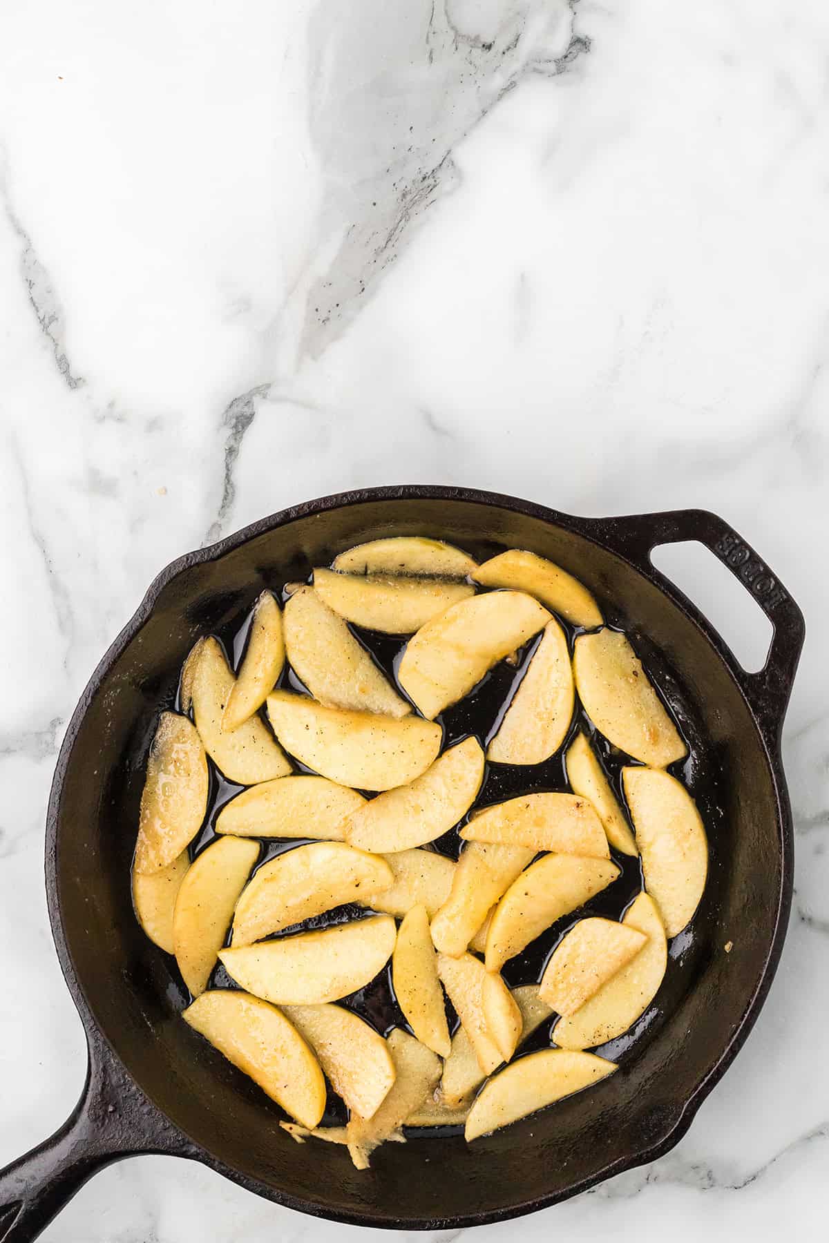 Sliced apples cooking in a black cast iron skillet on a white marble surface.