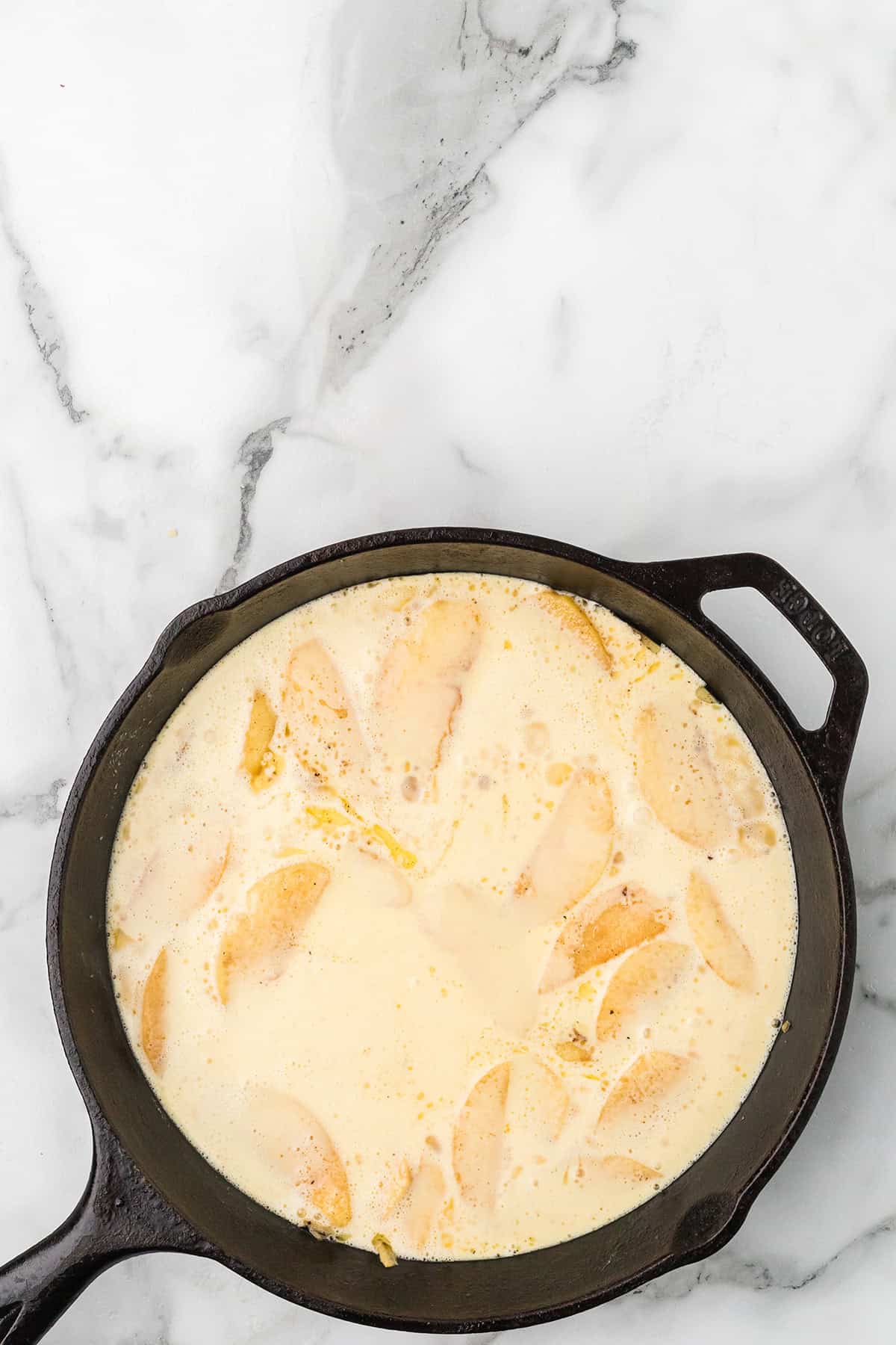 Sliced apples in creamy batter in a cast iron skillet on a white marble surface.