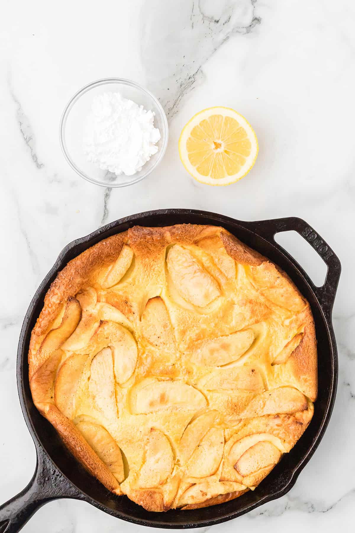 A cast iron skillet with apple pancake, next to a lemon half and a bowl of powdered sugar on marble.