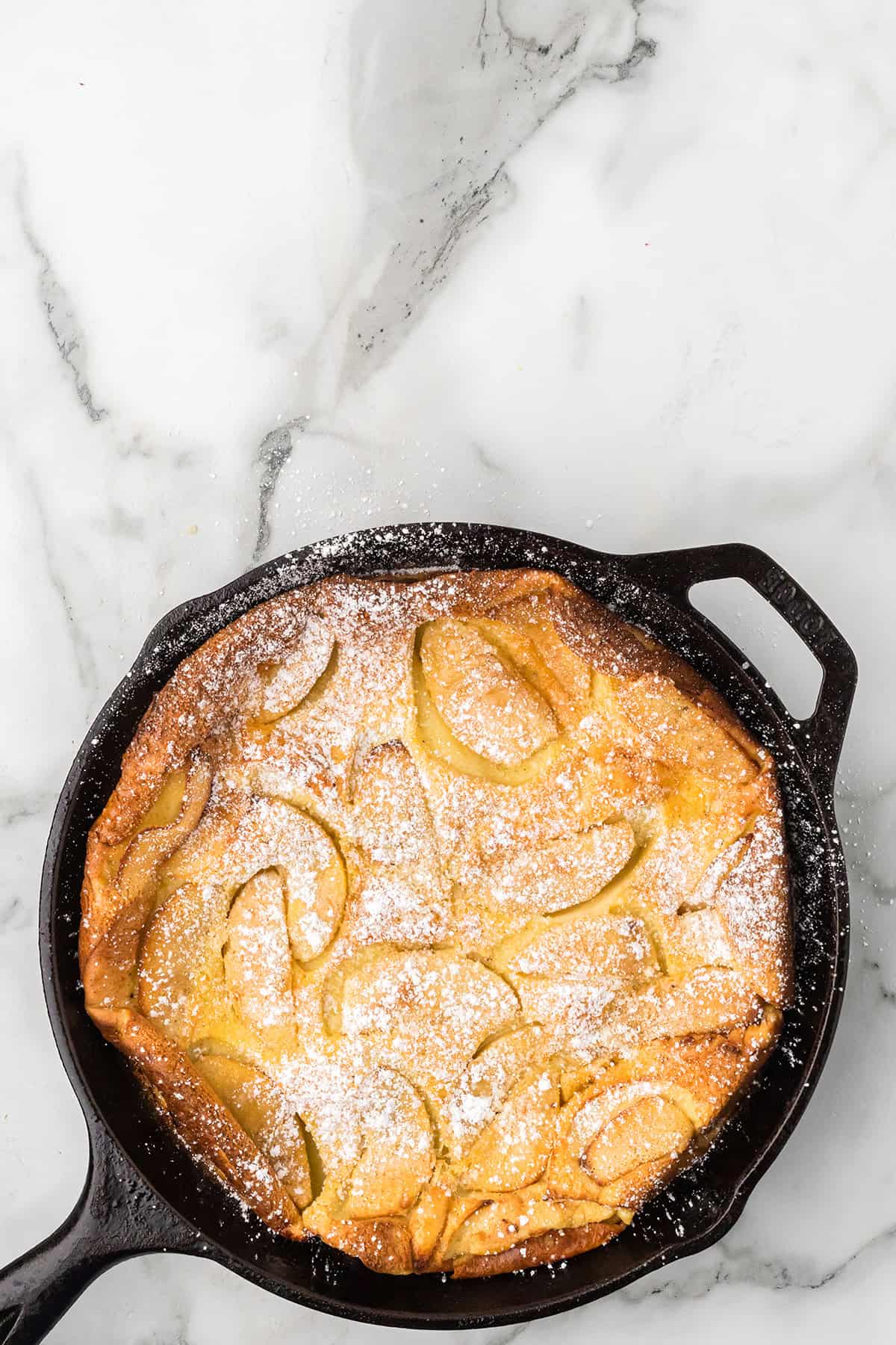 A baked apple Dutch baby pancake in a cast iron skillet, dusted with powdered sugar, on a marble surface.