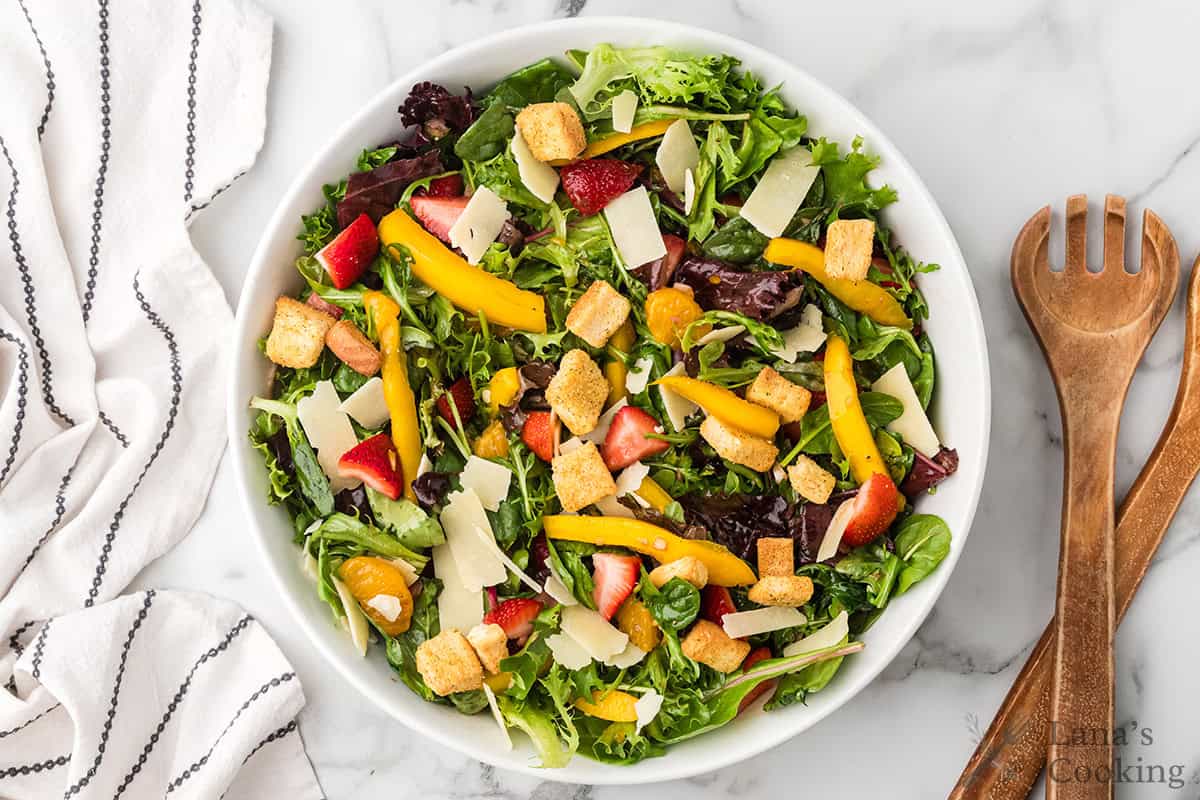 Fresh salad with mixed greens, strawberries, yellow tomatoes, croutons, and cheese shavings in a white bowl.