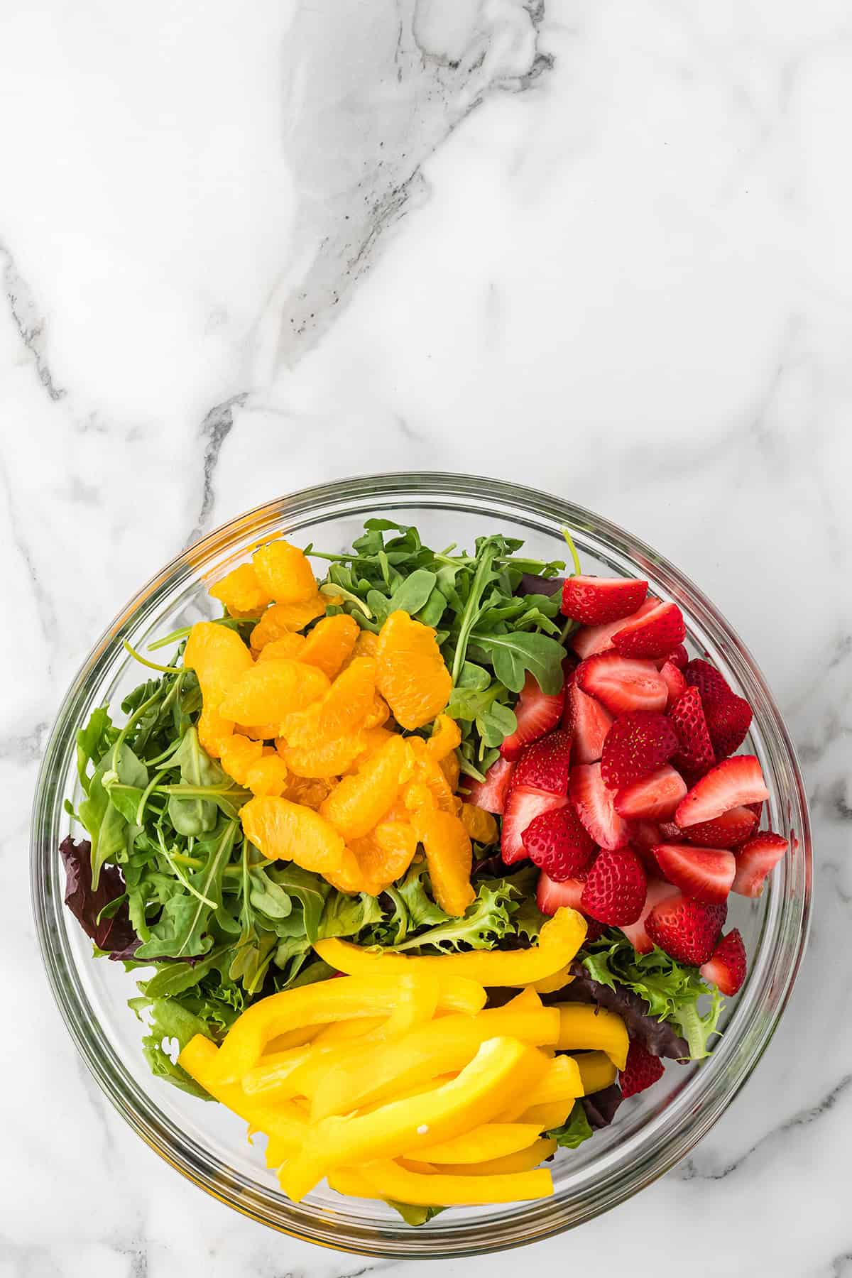 Glass bowl with leafy greens, sliced strawberries, orange segments, and yellow bell pepper strips on marble.