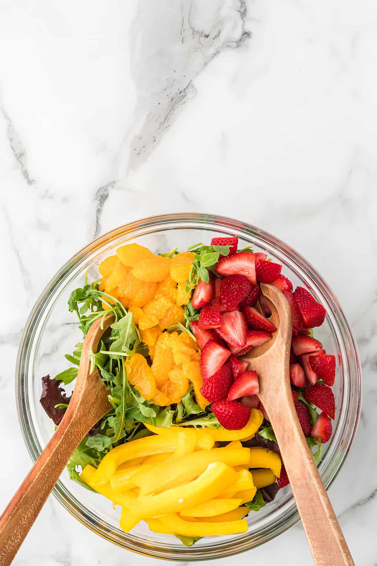 Glass bowl with arugula, orange slices, strawberries, and yellow bell pepper, with two wooden salad servers.