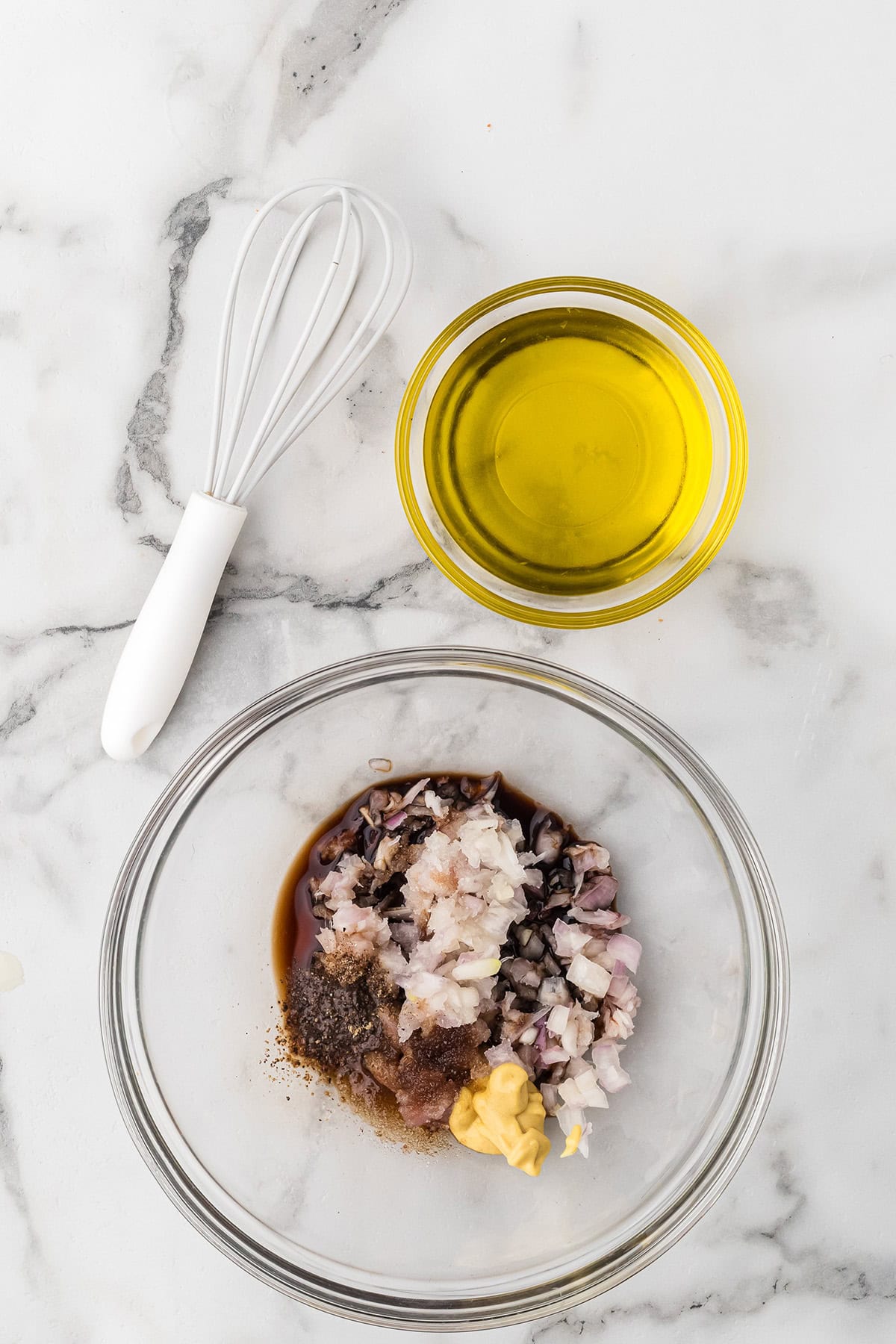 A whisk, a bowl of olive oil, and a bowl with chopped shallots, mustard, pepper, and balsamic vinegar on marble.