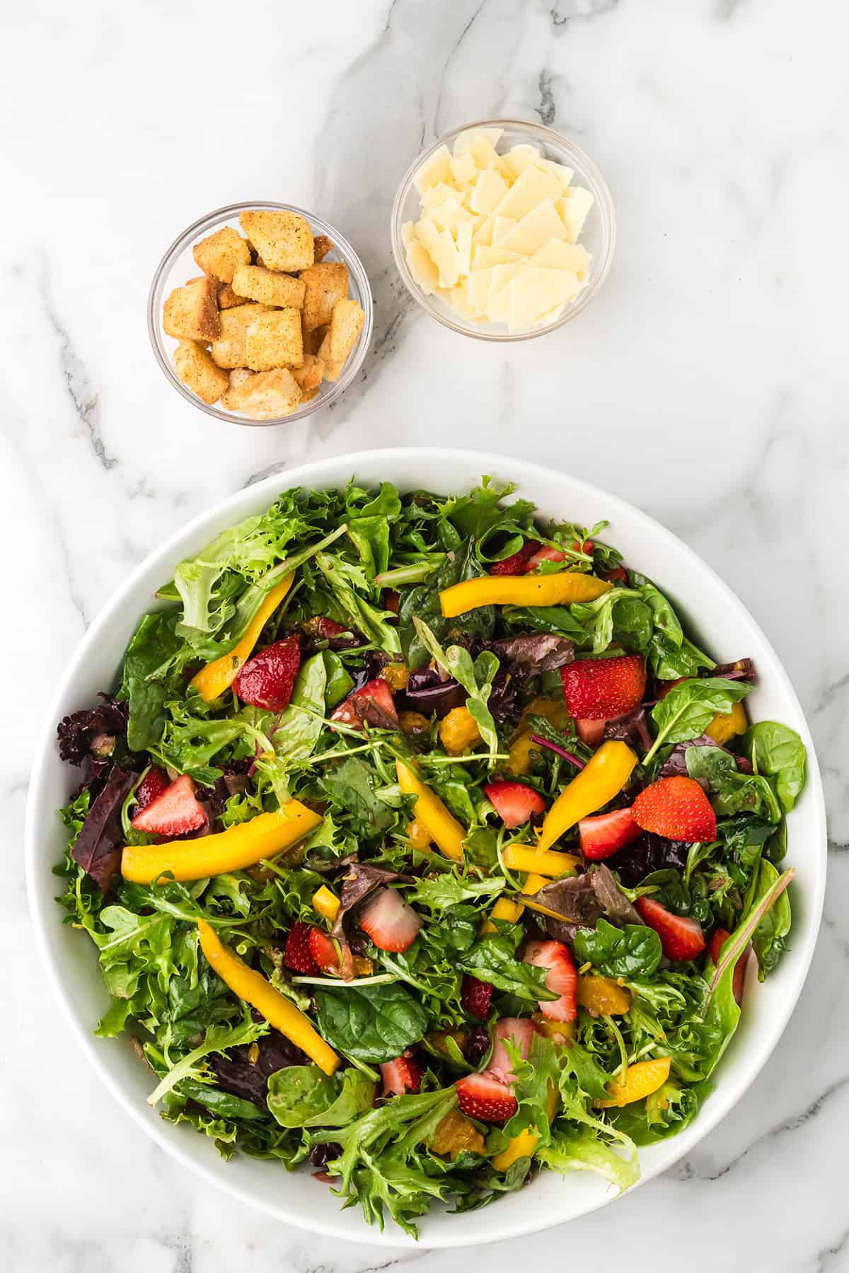 A bowl of mixed green salad with strawberries and yellow peppers, next to cups of croutons and cheese slices.
