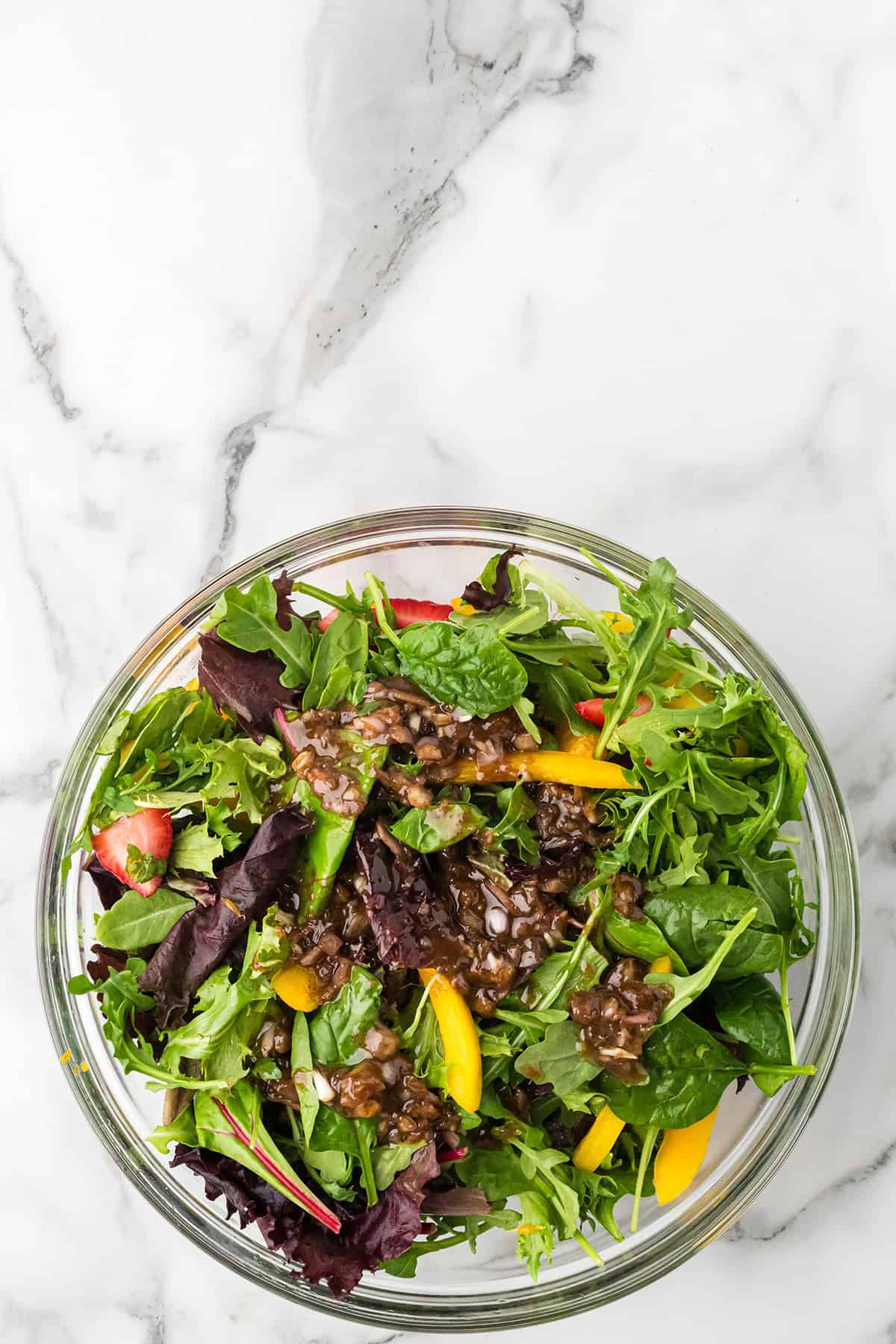A glass bowl of mixed green salad with yellow bell peppers and vinaigrette on a marble surface.