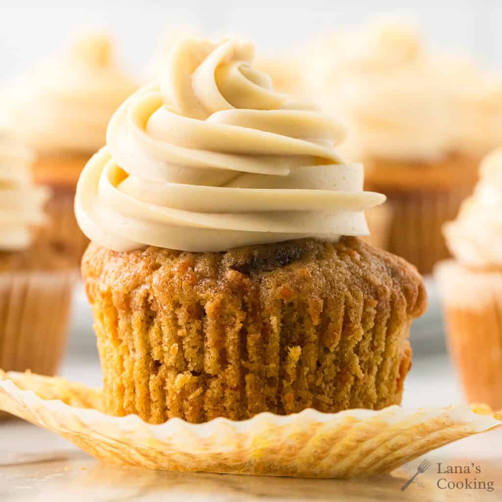 A carrot cake cupcake with swirled frosting, unwrapped from its liner, sits on a white surface.