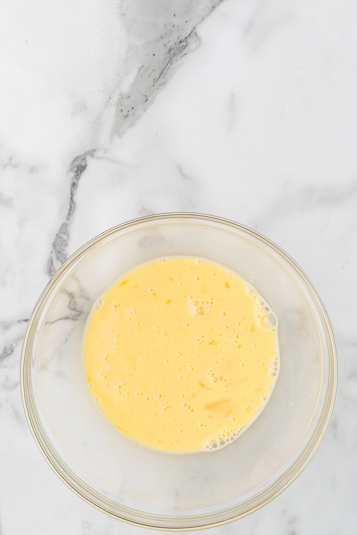 Glass bowl with beaten eggs and milk on a white marble surface.