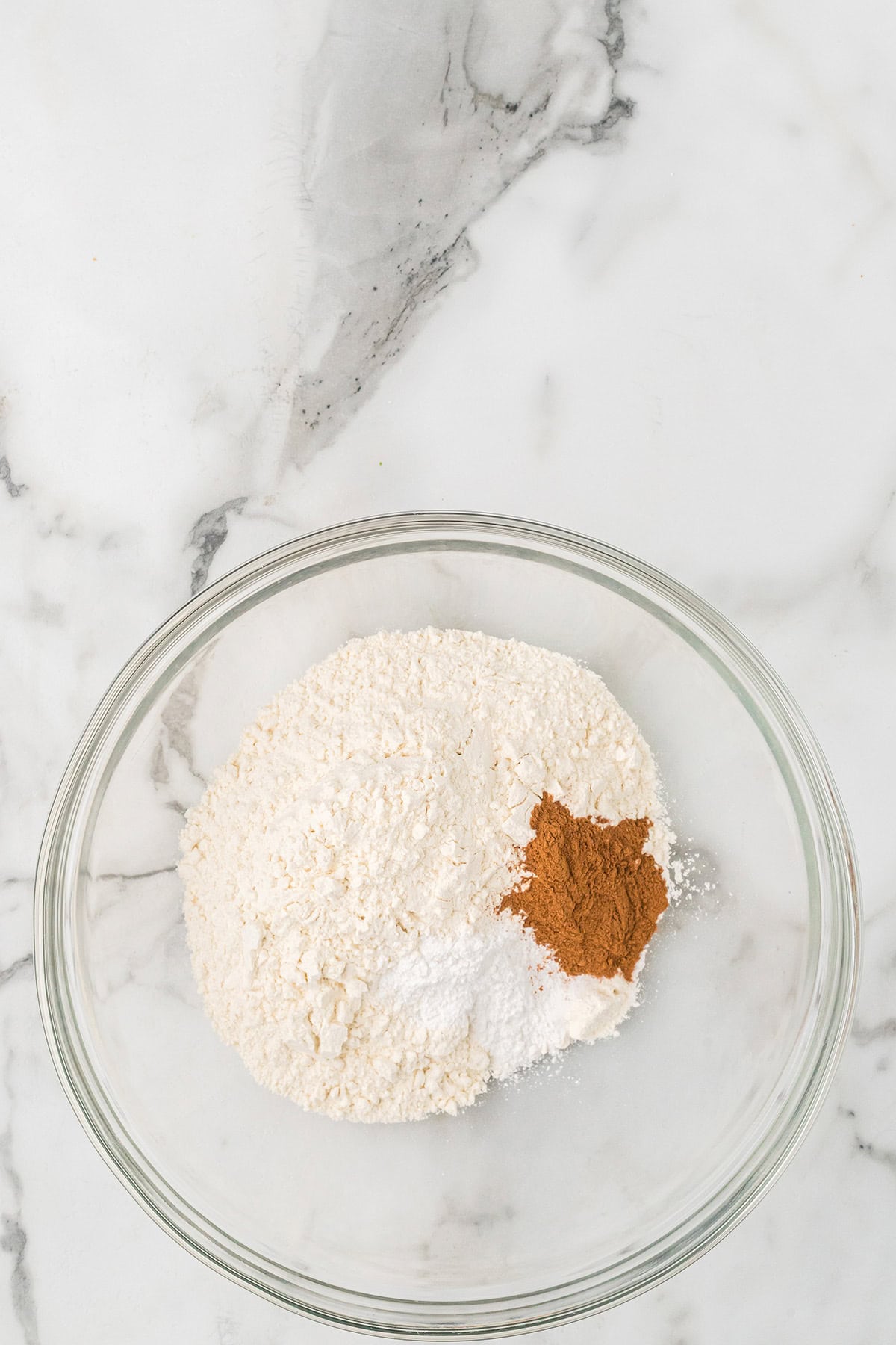 A glass bowl with flour, baking soda, and spices on a white marble surface.