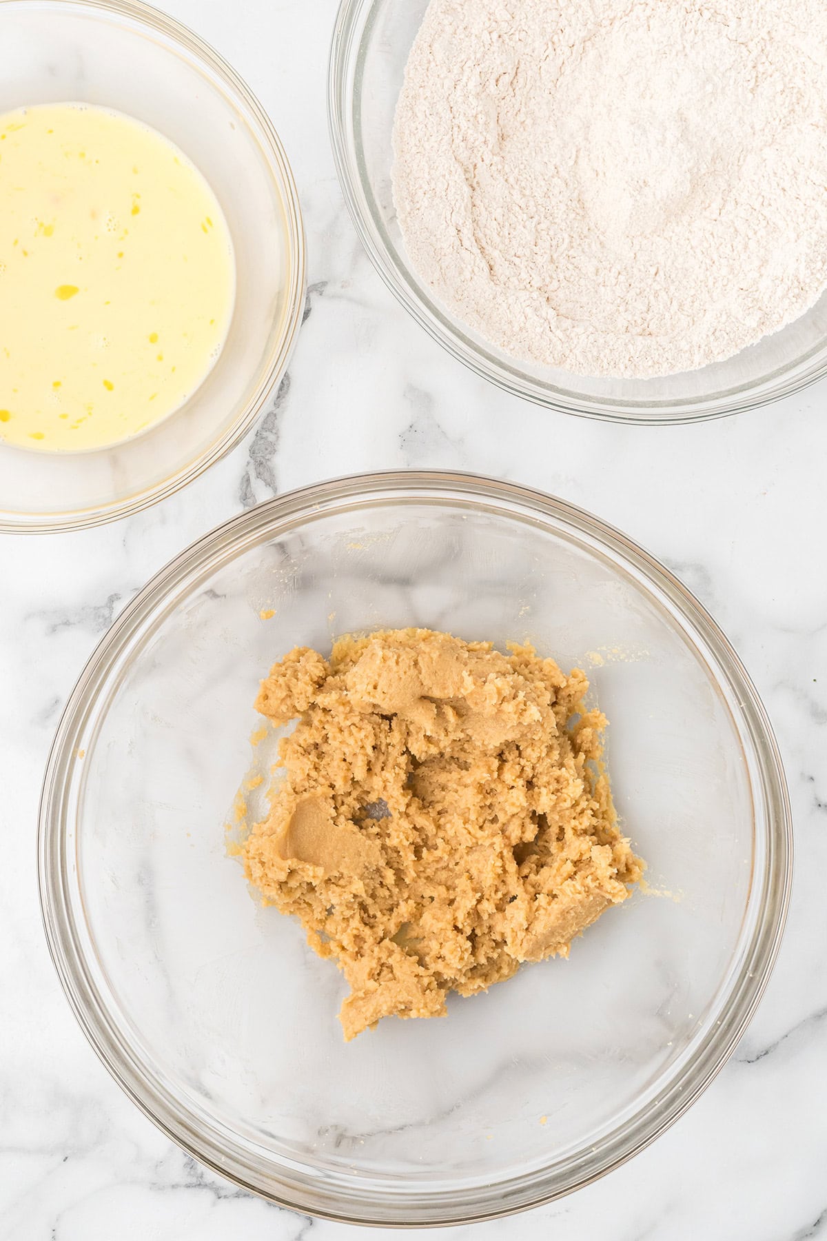 Three glass bowls with wet ingredients, dry ingredients, and brown sugar mixture on a marble surface.