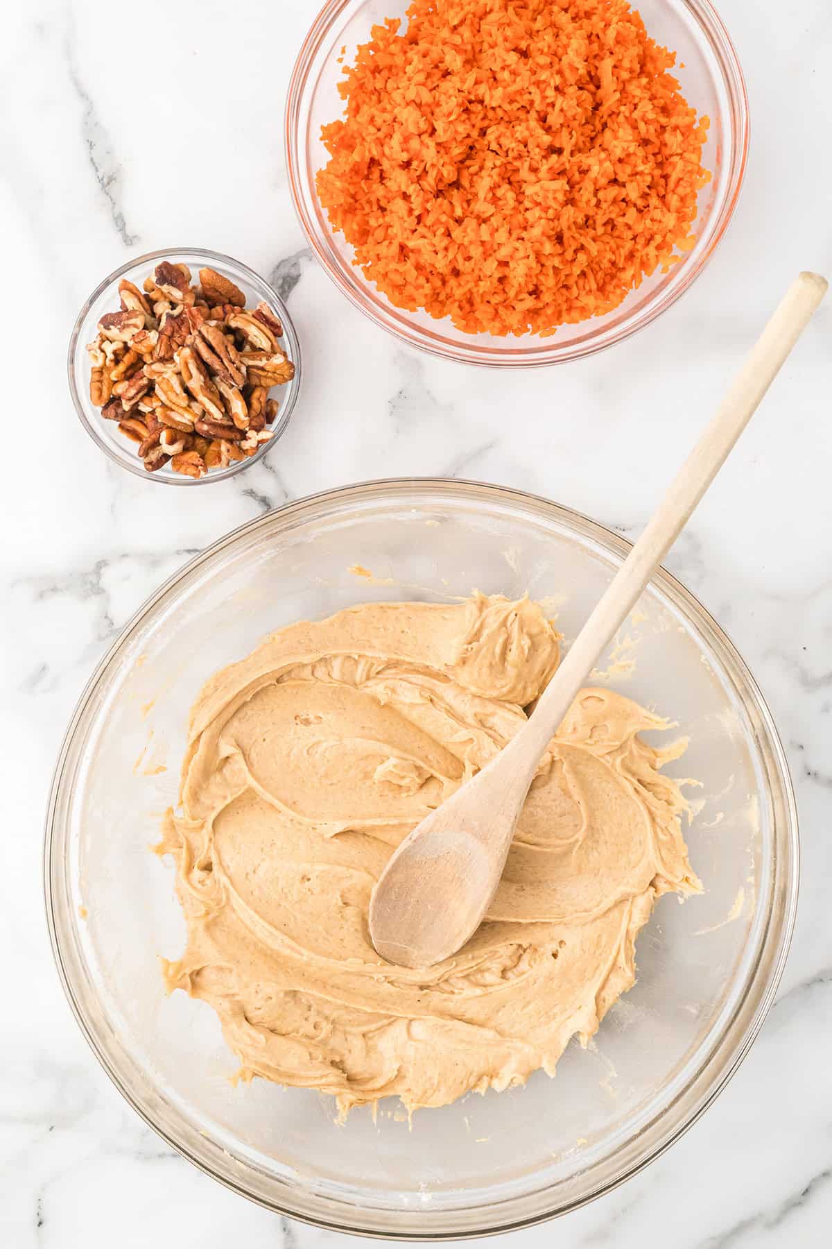 Large bowl of creamy batter with a wooden spoon, near bowls of chopped carrots and pecans on a marble counter.