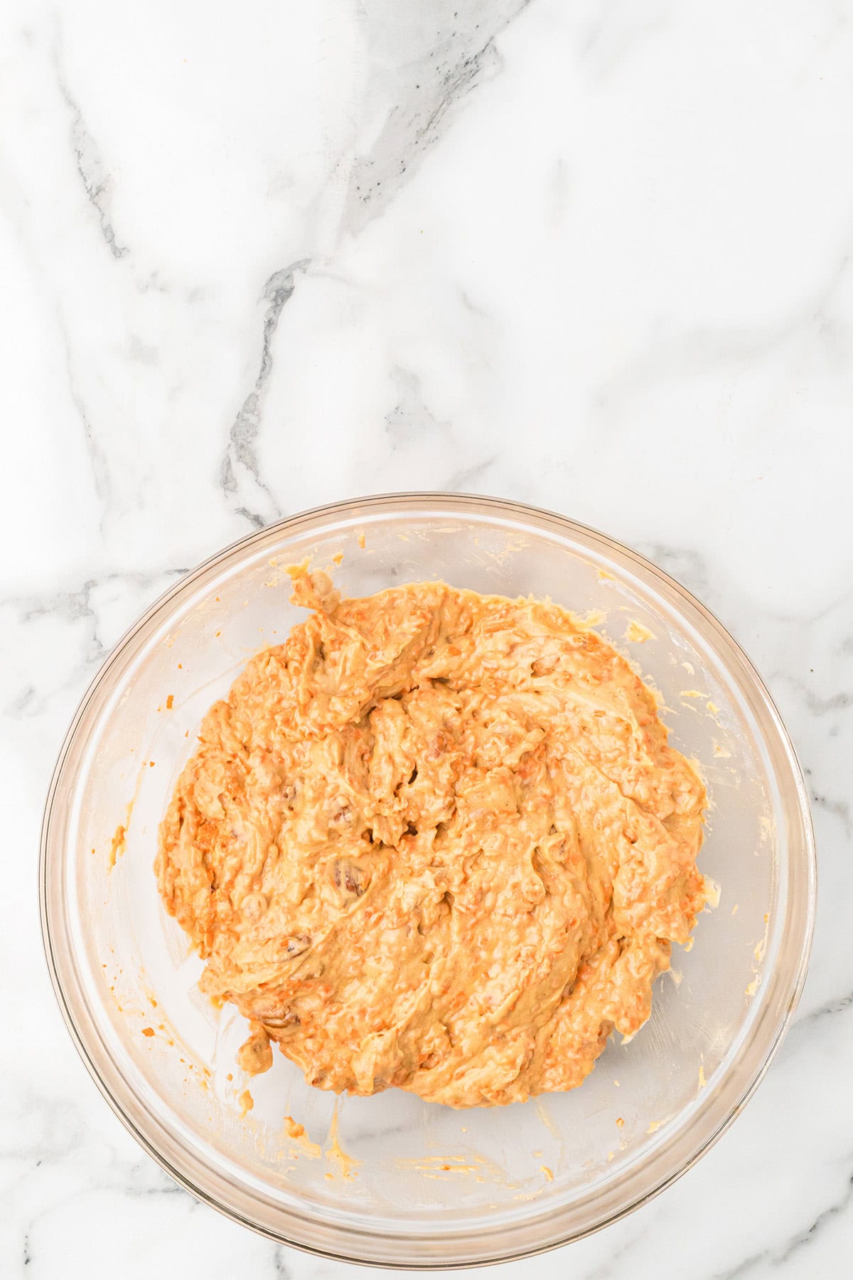 A glass bowl with carrot cake cupcake mixture on a white marble surface.