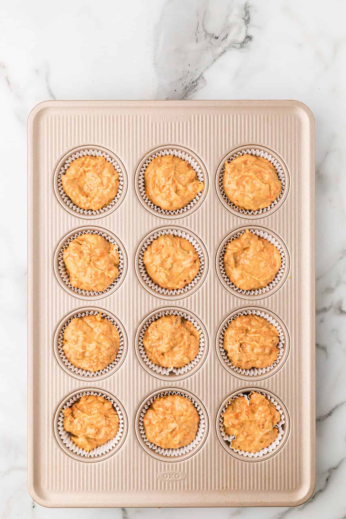 A cupcake tin with 12 carrot cake cupcakes ready to bake in paper liners on a marble countertop.