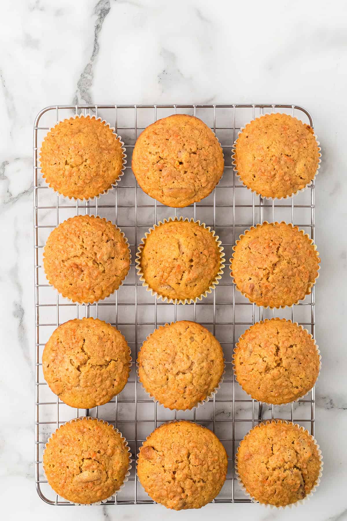 Twelve golden-brown cupcakes on a cooling rack, set on a white marble surface.