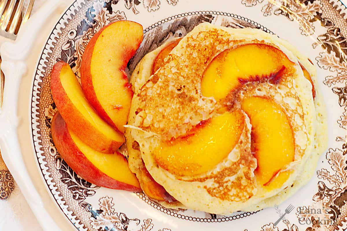 Pancakes with peach slices on a decorative plate, served with extra fresh peach slices on the side.