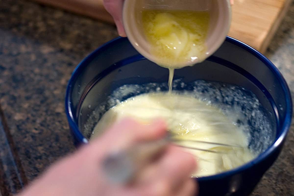 A person pours melted butter into a bowl of batter while whisking the mixture on a kitchen counter.