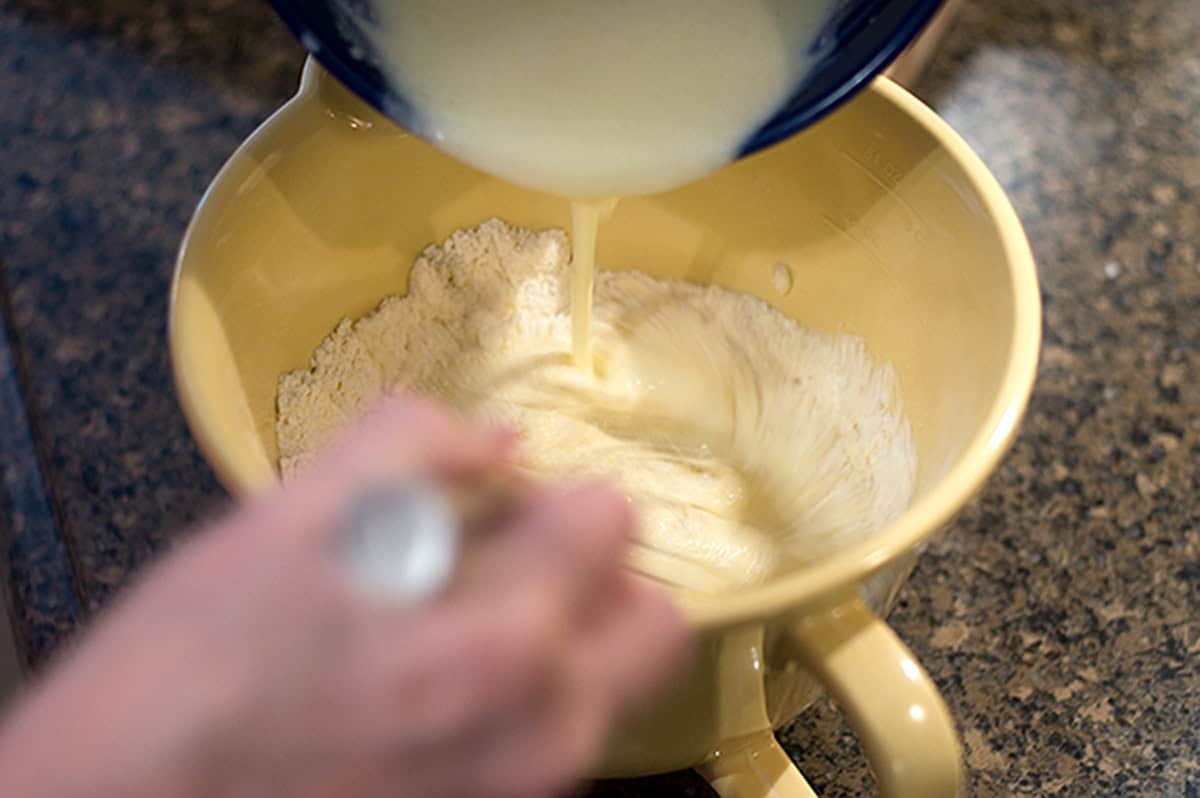 A person pours liquid into a bowl of flour and stirs the mixture with a spoon on a countertop.