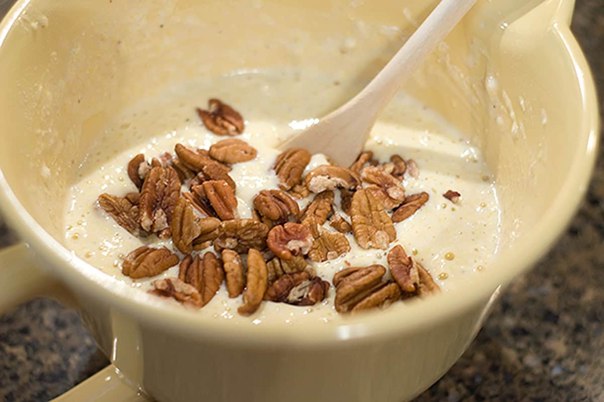 A mixing bowl with batter, pecan halves, and a wooden spoon on a kitchen counter.