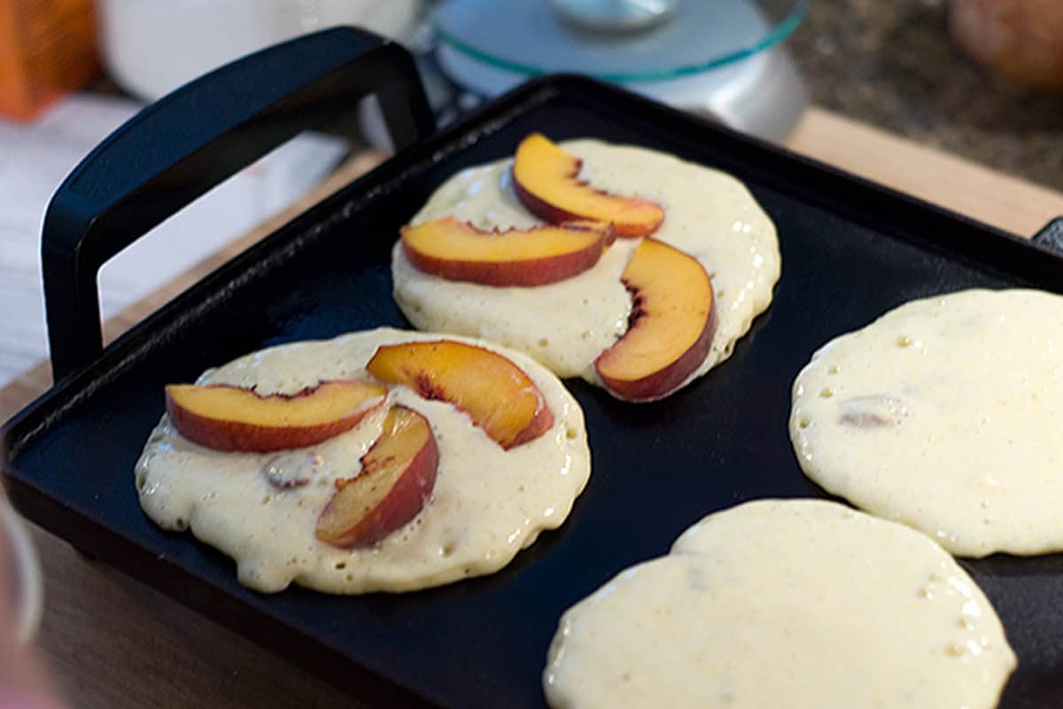 Three pancake batters on a griddle; two topped with fresh peach slices, one plain, cooking.
