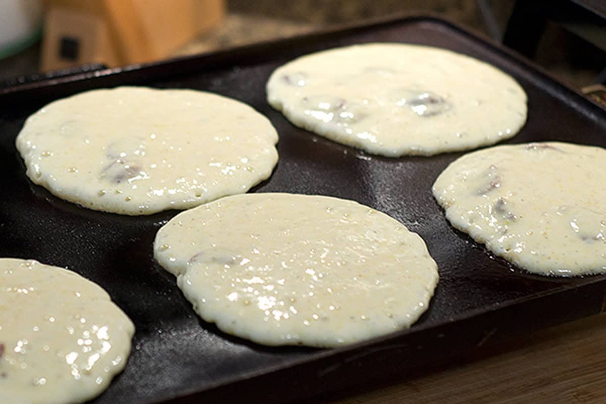 Five pancake batters cooking on a griddle, starting to bubble but not yet flipped.