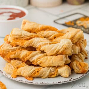 A plate of golden, twisted cheese straws with a cup of red dipping sauce in the background.