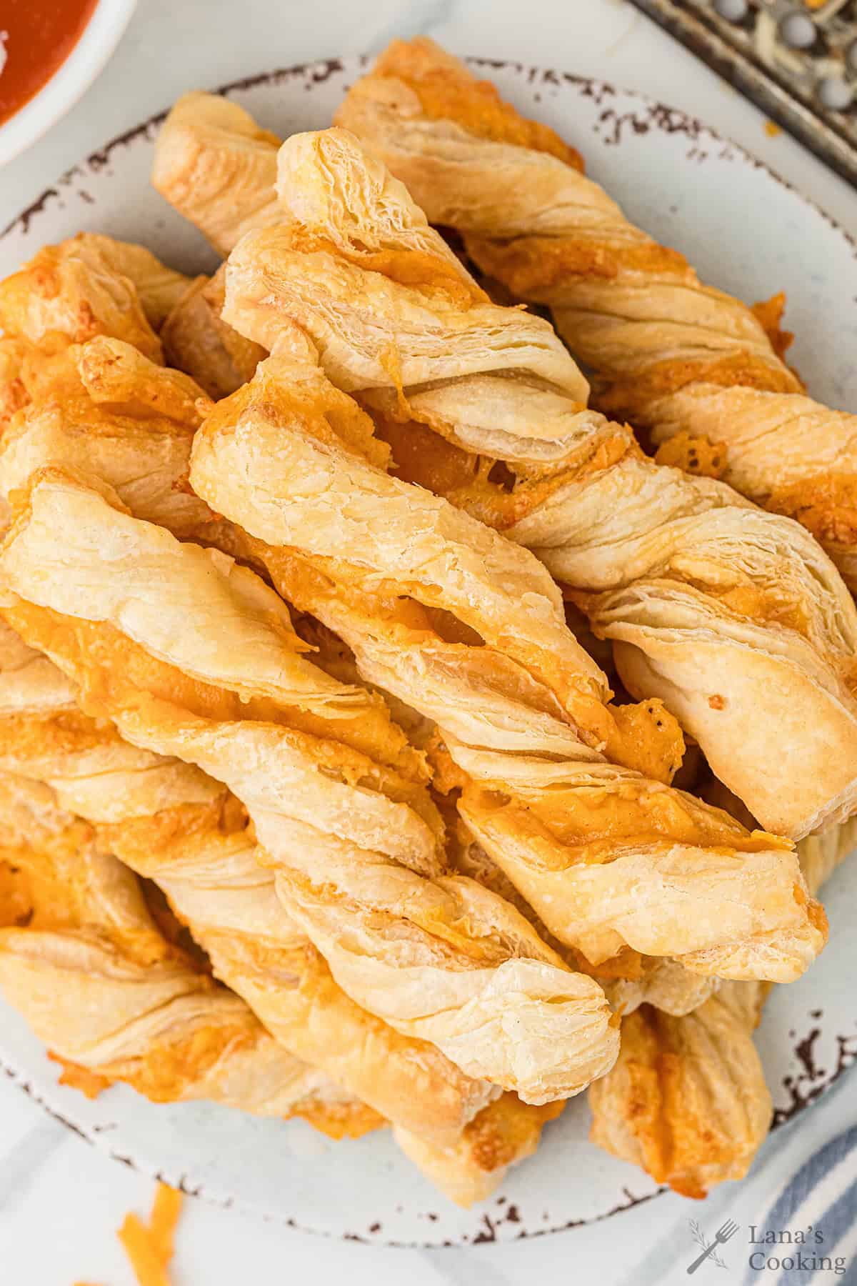 A plate of golden, twisted cheese straws made from puff pastry, stacked on a white plate.