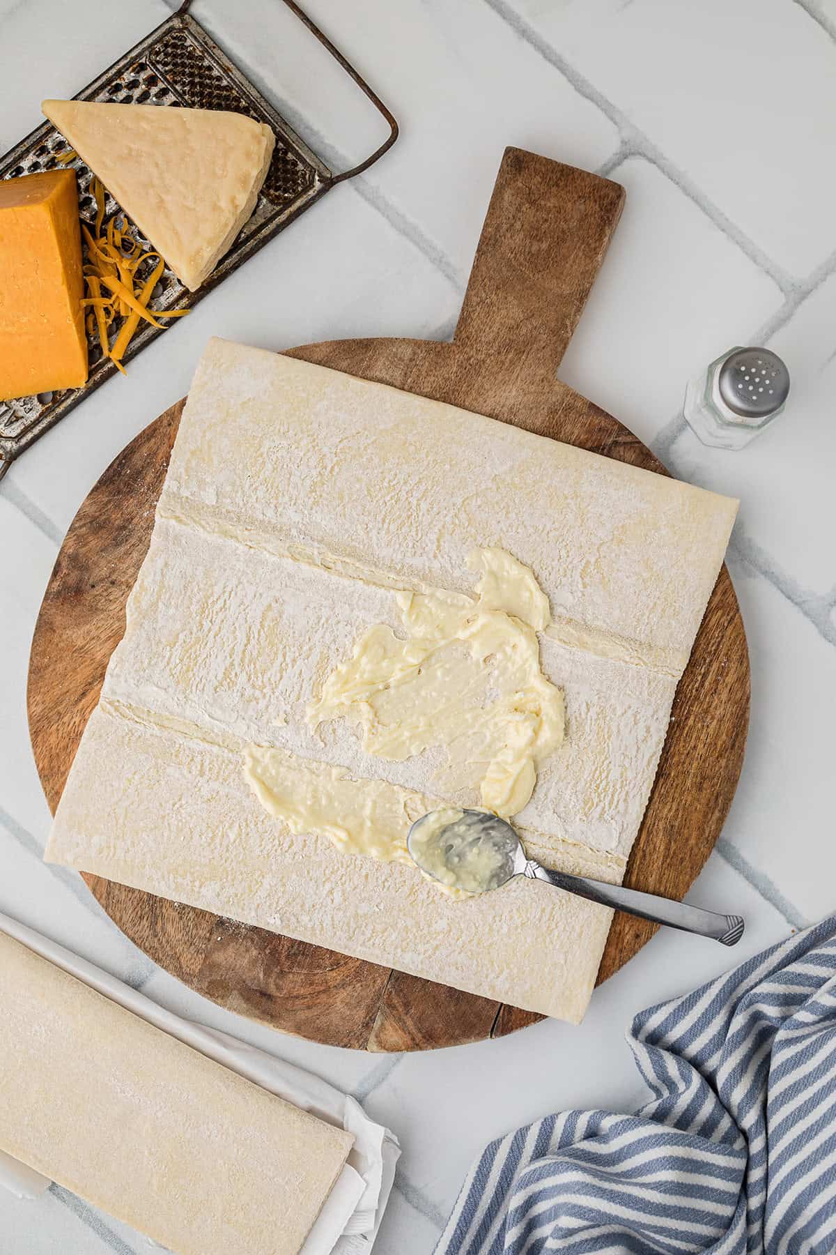 Puff pastry sheet on a wooden board, spread with butter; cheese and salt nearby on a white countertop.