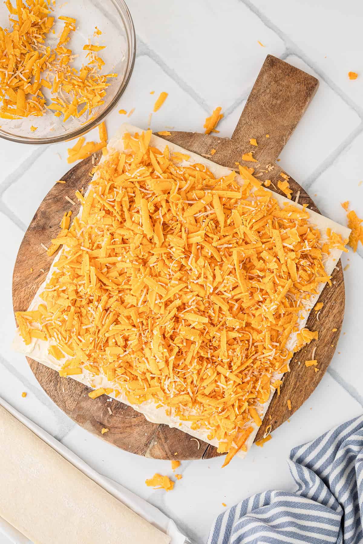 Square pastry topped with shredded cheddar cheese on a wooden board, with extra cheese in a bowl nearby.