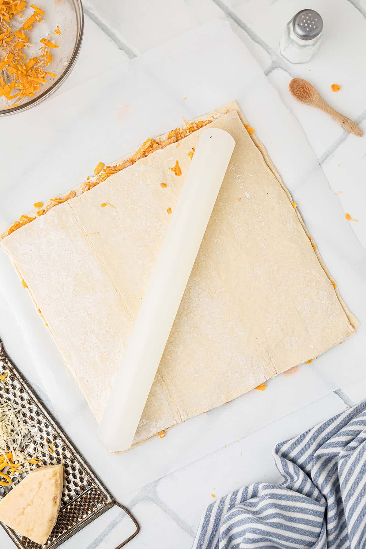 Sheet of puff pastry sprinkled with cheese being rolled out on a marble surface with a rolling pin.