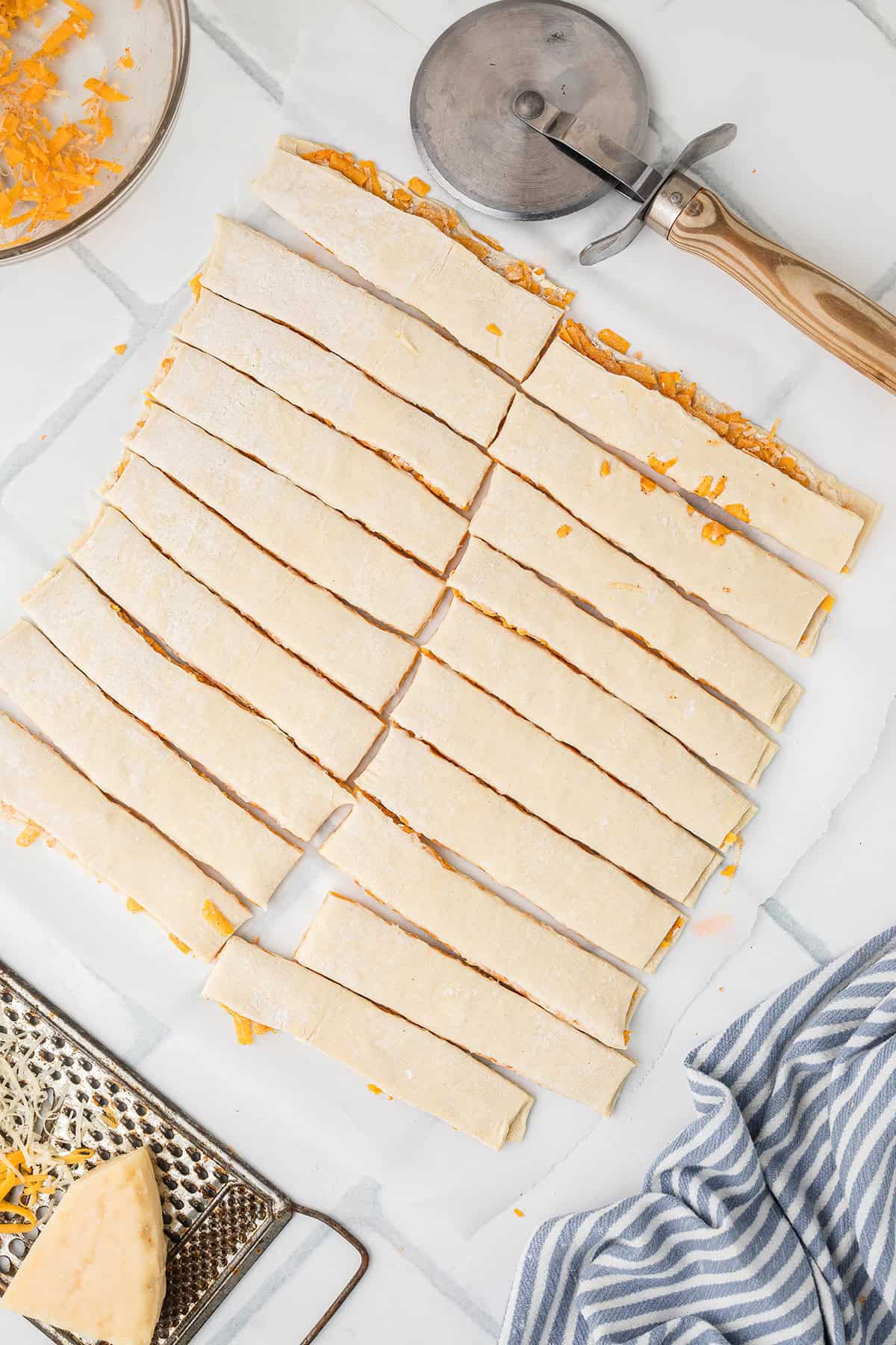 Unbaked dough strips with cheese, a pizza cutter, and a cheese grater on a marble surface.