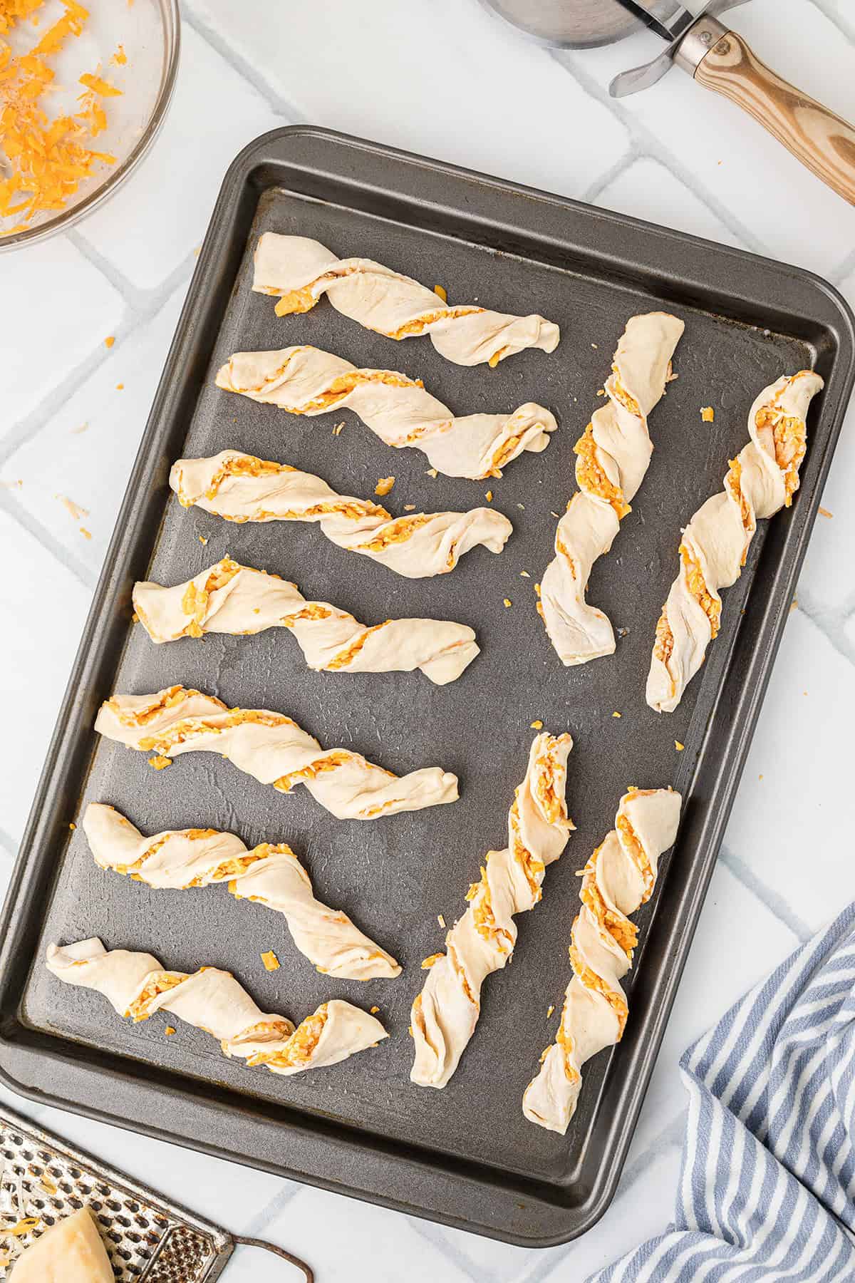 Unbaked twisted cheese straws arranged on a dark baking sheet, ready for the oven.
