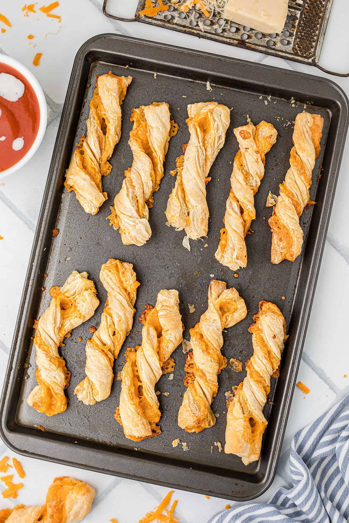 Baked cheese straws arranged on a dark baking sheet, with grated cheese and sauce nearby.