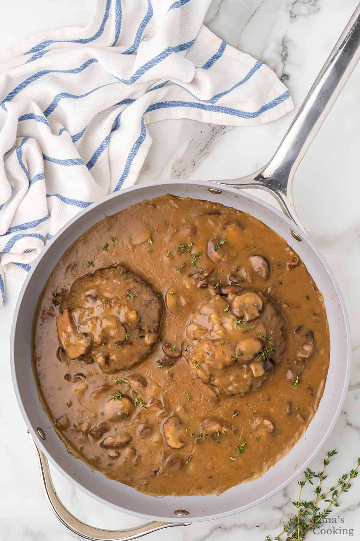 A skillet with two Salisbury steaks in mushroom gravy, garnished with herbs, beside a striped towel on marble.