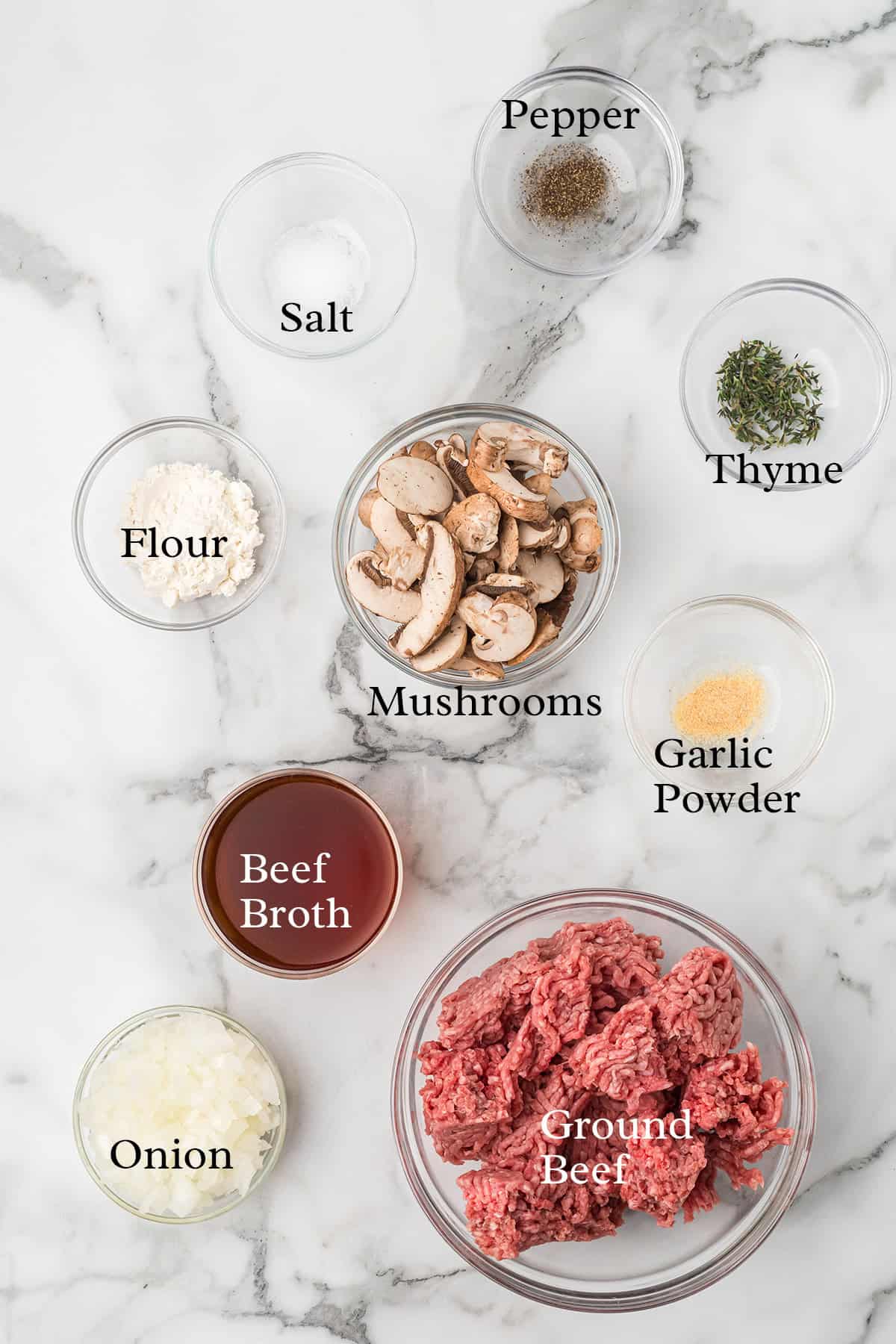 Bowls with ground beef, beef broth, mushrooms, onions, flour, salt, pepper, thyme, and garlic powder on marble.