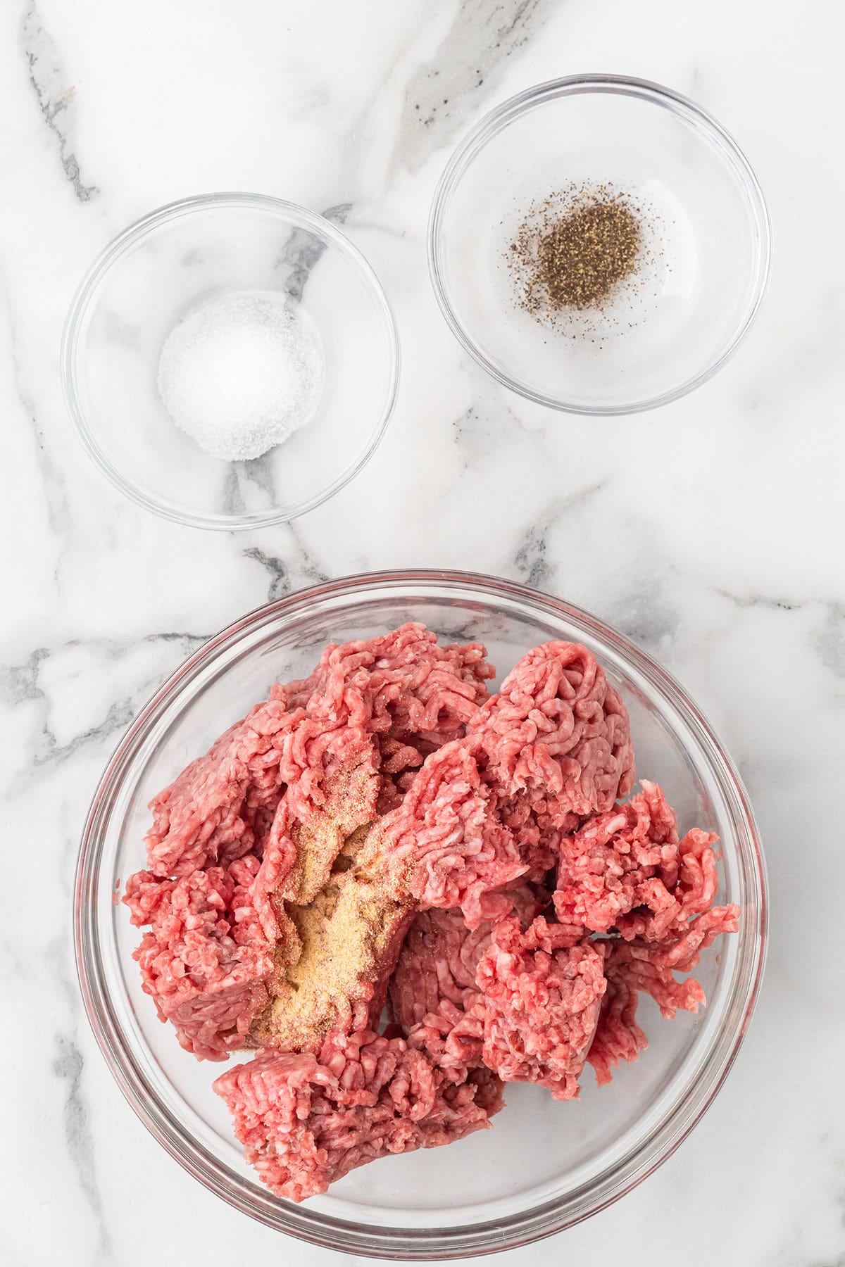 Ground beef in a glass bowl with seasonings, next to small bowls of salt and pepper, on a marble surface.
