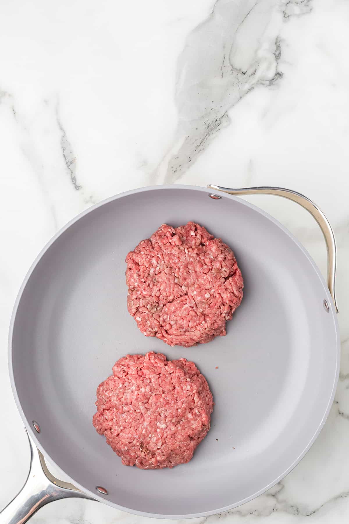 Two raw ground beef patties in a gray frying pan on a white marble countertop.