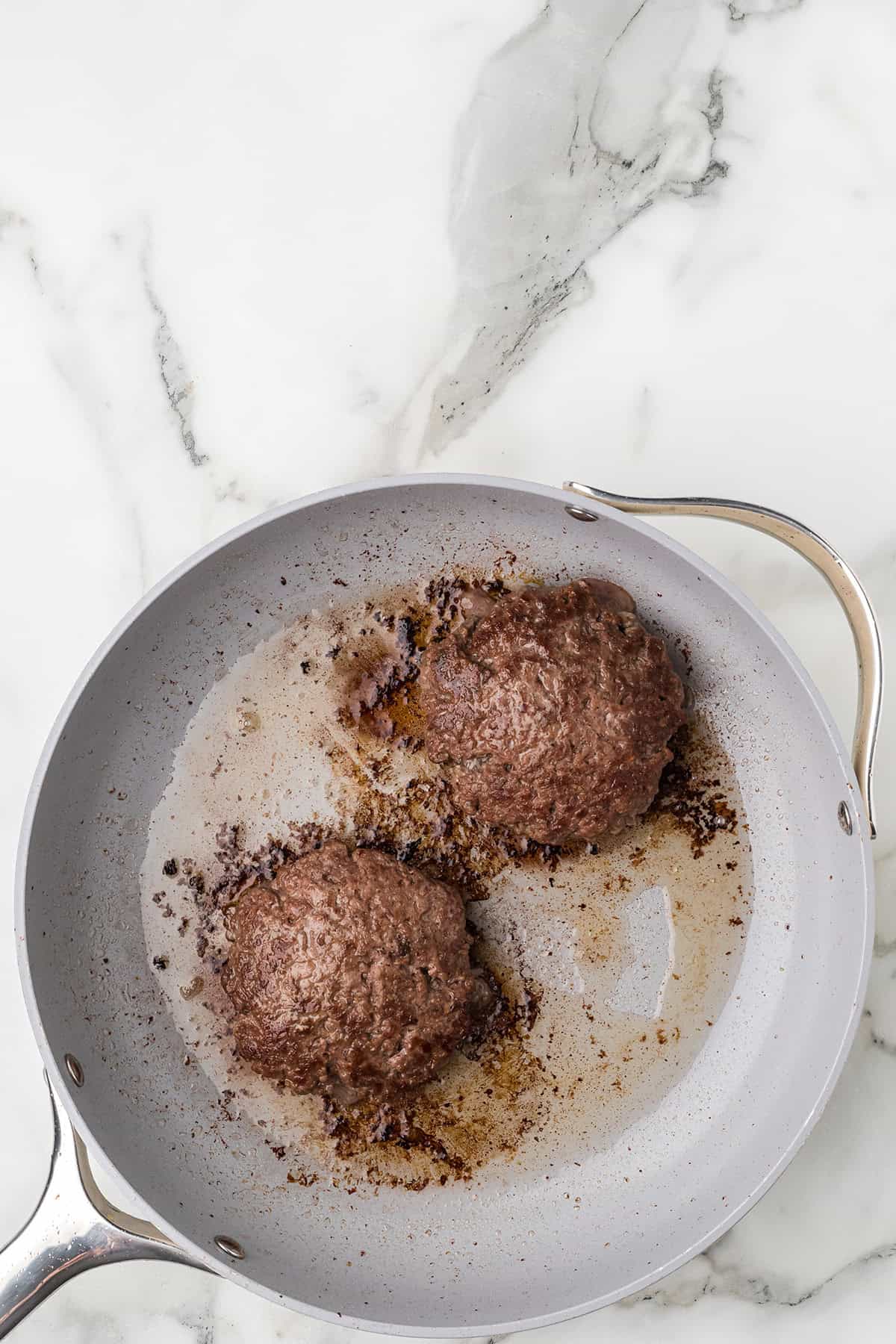 Two cooked burger patties in a frying pan with browned bits and oil, on a marble countertop.