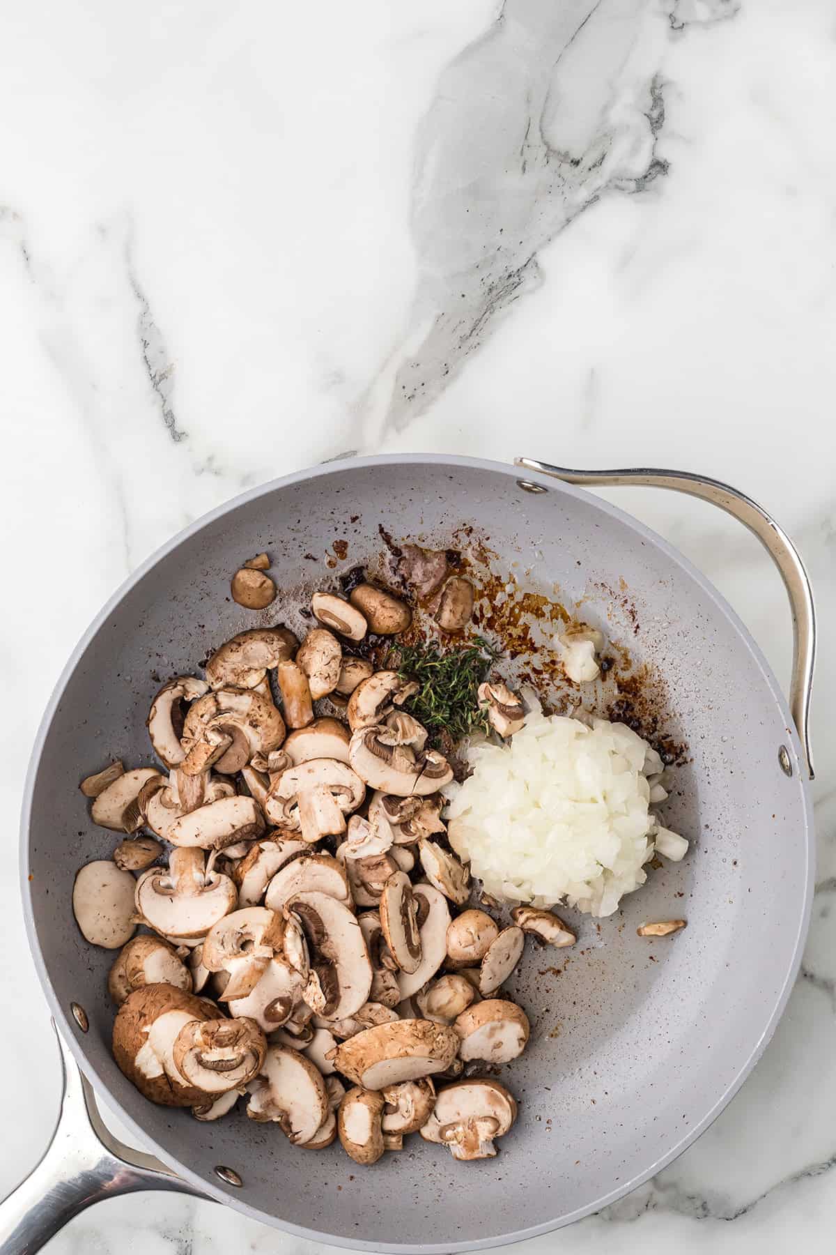 Sliced mushrooms and chopped onions cooking in a gray skillet on a marble countertop.