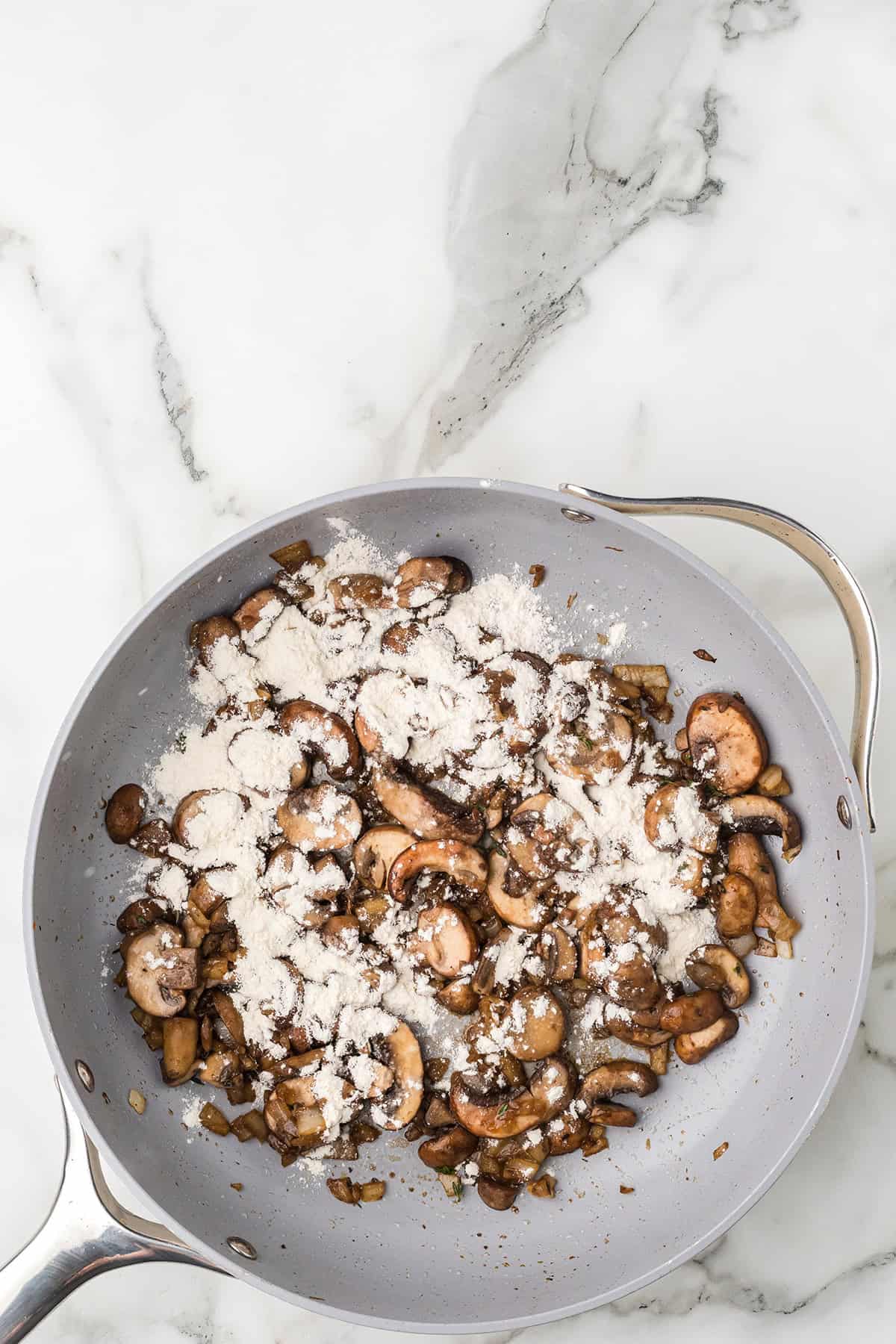 Sliced mushrooms cooking in a skillet with a sprinkle of flour on top, on a marble countertop.